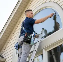 Professional window cleaner on a ladder using a squeegee to clean an exterior arched window, Tucson, AZ.